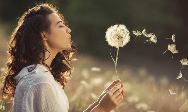 Woman Outdoors Blowing Dandelion Seeds. Symbolizing Airborne Pollen and Seasonal Allergies that can Trigger Post Nasal Drip.